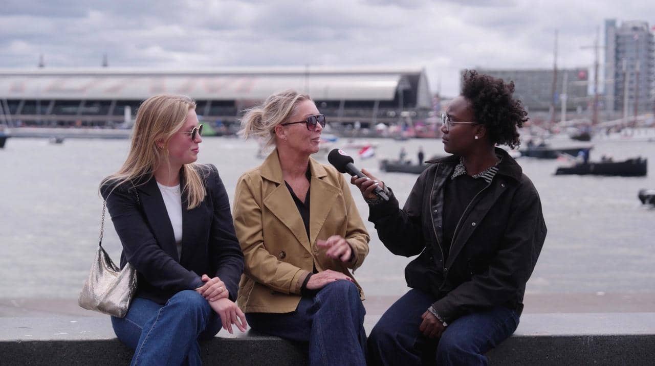 Drie vrouwen zitten op de rand van een boot met op de achtergrond 't IJ en het Centraal Station van Amsterdam. Een van de vrouwen houdt een microfoon voor een andere vrouw.