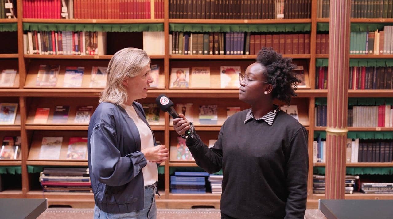Twee vrouwen staan in het Rijksmuseum in een zaal met boeken op de achtergrond. Een van de vrouwen houdt een microfoon vast terwijl de ander praat.